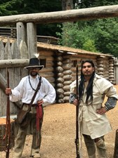 Zachary Stocks (left)  and Izzy Sanchez (right) stand infront of Fort Clatsop dressed in period attire with flintlock  rifles.