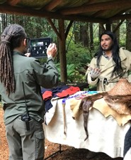 Park Ranger Zachary Stocks films Park Ranger Izzy Sanchez on an Ipad for a class. Glass beads, military uniform coats, and furs cover the table.
